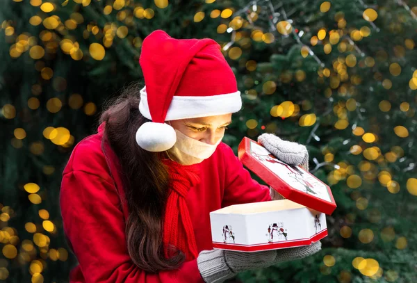Kırmızı Noel Baba şapkalı ve beyaz tıbbi koruyucu maskeli tatlı bir Avrupalı kız Noel hediyesine bakar ve mutlu olur. Coronavirus Yeni Yıl ve Noel kendini izole edip karantinaya alındı.