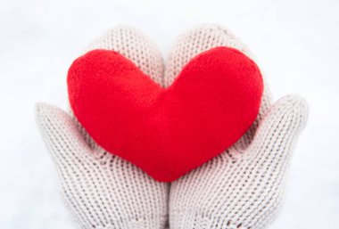 Bej eldivenli kumaştan yapılmış kırmızı yumuşak kalp oyuncağını yeni yağmış beyaz karın arka planında tut. Sevgililer Günü için yaratıcı tebrik kartı.