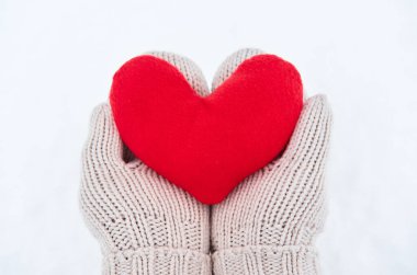 Bej eldivenli kumaştan yapılmış kırmızı yumuşak kalp oyuncağını yeni yağmış beyaz karın arka planında tut. Sevgililer Günü için yaratıcı tebrik kartı.