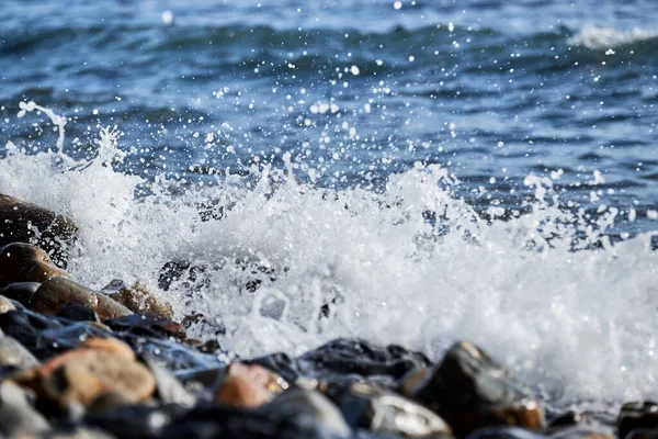 Small wave rolls onto shore and splash of foam flies in different directions. Beautiful marine minimalist screensaver. Sea waves break on shingle beach.