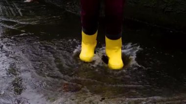 Sarı lastik çizmelerle su birikintilerinden atla. Yağmurda yürümek ve kötü havanın tadını çıkarmak çocukluğa düşmek ve ıslak su birikintilerinde koşmak. Yüzü olmayan bacak videosu.