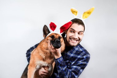 Sarı tavşan kulaklı beyaz adam, kırmızı kabarık kulaklı Alman çoban köpeğine sarılıyor. Paskalya kutlamaları için yaratıcı bir bayrak. Beyaz arka planda insan ve köpek. Tavşan kulaklı çoban köpeği..