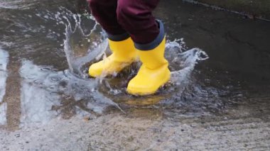Sarı lastik çizmelerle su birikintilerinden atla. Yağmurda yürümek ve kötü havanın tadını çıkarmak çocukluğa düşmek ve ıslak su birikintilerinde koşmak. Yüzü olmayan bacak videosu.