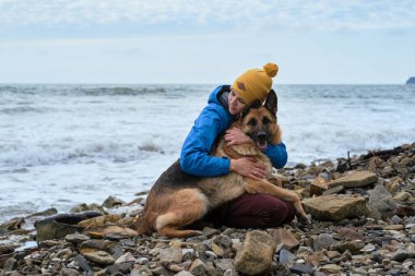 Çoban köpeğine iki elinle sıkıca sarıl. Genç, beyaz bir kadın boş zamanlarını köpekle sahilde oturarak geçiriyor. Kız ve Alman çoban köpeği temiz havada. Sahibi ve safkan köpeği sevmek.