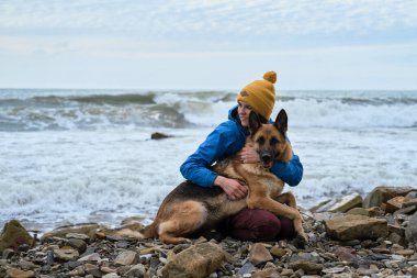 Çoban köpeğine iki elinle sıkıca sarıl. Genç, beyaz bir kadın boş zamanlarını köpekle sahilde oturarak geçiriyor. Kız ve Alman çoban köpeği temiz havada. Sahibi ve safkan köpeği sevmek.