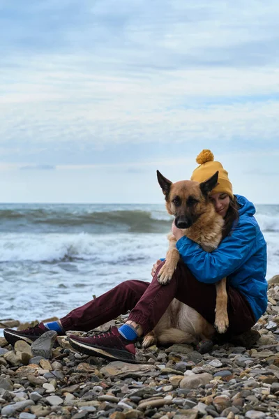 Çoban köpeğine iki elinle sıkıca sarıl. Genç, beyaz bir kadın boş zamanlarını köpekle sahilde oturarak geçiriyor. Kız ve Alman çoban köpeği temiz havada. Sahibi ve safkan köpeği sevmek.