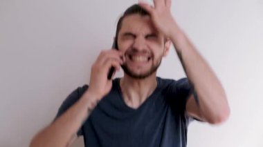 Beyaz arka planda mavi tişörtlü, sakallı ve şık saç stili genç beyaz adam telefonda konuşuyor, kızgın ve ellerini sallıyor. Adam tatsız bir konuşmadan sonra akıllı telefonu bir kenara atıyor..