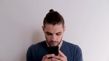 Mavi sakallı, beyaz tişörtlü genç beyaz adam beyaz duvara karşı akıllı telefon kullanarak internette mesaj yazıyor. İnternet 'te iş yazışmaları.