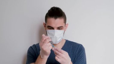 Mavi tişörtlü, tıbbi koruyucu maskeli, beyaz lateks eldivenli beyaz arka planda duran ve şırıngayı ilaçla dolduran beyaz bir beyaz öğrenci. Adam iğneyi yapacak..