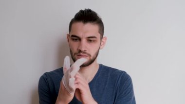 Beyaz arka plandaki mavi tişörtlü yakışıklı beyaz adam kendini çeşitli virüs ve bakterilerden korumak için lateks şeffaf eldivenler giyer. Kişisel koruma, 4k video.