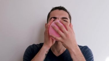 Beyaz arka planda mavi tişörtlü, sakallı ve at kuyruklu hoş beyaz adam ağzıyla pembe balonu şişiriyor. Balonu şişir ve bayramı kutla.