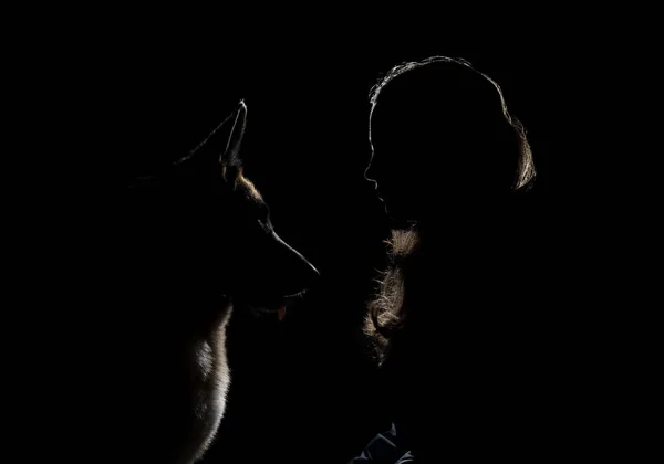 Alman çoban köpeği ve sahibinin profili oturup birbirine bakar. Kız ve evcil hayvan siluetleri çok güzel çizilmiş. Siyah arka plandaki parlak ışıkla aydınlatılmış köpek ve kadın silueti..