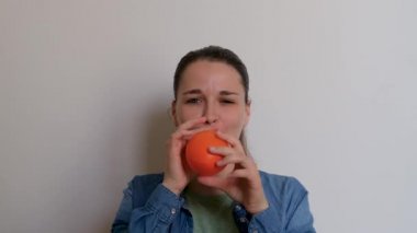 Young pretty woman of Caucasian European appearance inflates orange balloon with her mouth on white background. Girl prepares for holiday and inflates balloon and then lets it fly away.