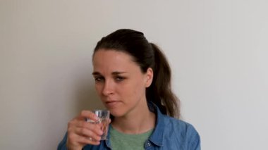 Young Caucasian European girl in shirt sits on white background and drinks pills with water. Treatment with medications and antibiotics at home. 4K footage of taking tablets by person.