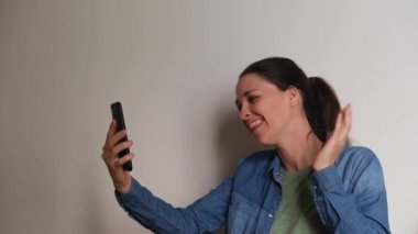 Young Caucasian woman on white background talks on video link using smartphone and looks at phone screen smiles rejoices and waves her hands. Important conversation over Internet online. 4K footage.