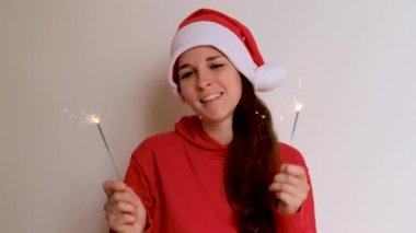 Parlak kapüşonlu ve kırmızı Noel Baba şapkalı beyaz arka planda dans eden ve iki maytap sallayan beyaz bir kız. Yeni yıl Noel 4K görüntüsü yüksek kaliteli.