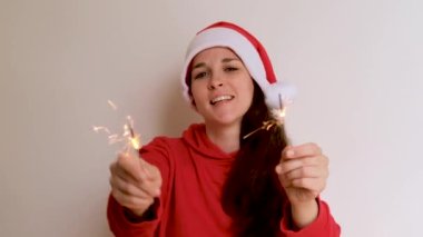 Parlak kapüşonlu ve kırmızı Noel Baba şapkalı beyaz arka planda dans eden ve iki maytap sallayan beyaz bir kız. Yeni yıl Noel 4K görüntüsü yüksek kaliteli.