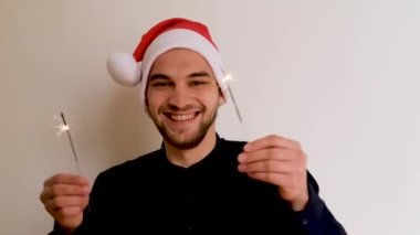 Gömleği sakallı, kırmızı Noel Baba şapkalı, beyaz arka planda dans eden ve havai fişekleri sağa sola sallayan yakışıklı beyaz bir adam. Yeni Yıl Noel 4K görüntüleri.
