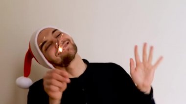 Gömleği sakallı, kırmızı Noel Baba şapkalı, beyaz arka planda dans eden ve havai fişekleri sağa sola sallayan yakışıklı beyaz bir adam. Yeni Yıl Noel 4K görüntüleri.
