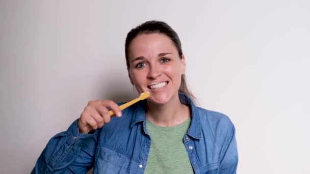 身穿蓝色衬衫的年轻可爱的白人女孩正在用黄色的牙刷用白色的背景刷牙 妇女每天都照顾口腔卫生 4K高质量镜头 — 图库视频影像