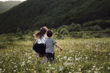 Çocuklar, erkek ve kız kardeşler papatya tarlasında neşeyle el ele koşuşturuyorlar. Açık alanda yürüyen sevimli çocuklar güzel çiçeklerin arasında rahatlar. Mutlu çocukluk doğası.