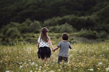 Çocuklar, erkek ve kız kardeşler papatya tarlasında neşeyle el ele koşuşturuyorlar. Açık alanda yürüyen sevimli çocuklar güzel çiçeklerin arasında rahatlar. Mutlu çocukluk doğası.
