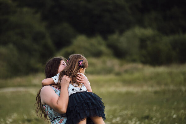 Young mother hugs her daughter tightly and she laughs with pleasure and embarrassment. Mother and child in chamomile field love each other. Beautiful family and joy of motherhood.