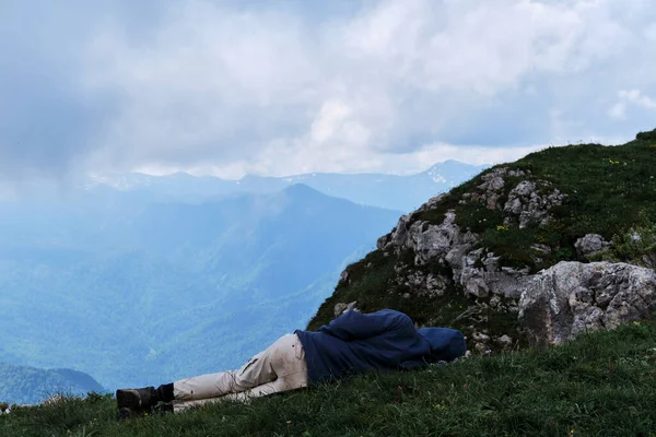 Male tourist fell asleep in grass on top of mountain against the background of low clouds and snow capped mountains in distance. Traveler was tired in campaign and decided to rest.