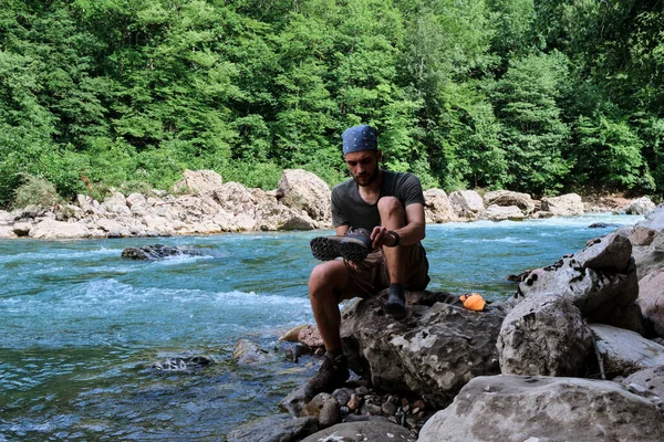 Getting legs wet in icy river. Young handsome Caucasian male traveler with bandana on his head and beard is sitting on stone near mountain river and takes off his shoes and socks to wash his feet.