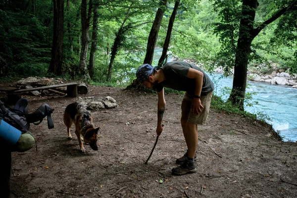 Young handsome Caucasian male traveler with bandana on his head and beard draws line on ground with stick to set up tent. German Shepherd traveler stands nearby and helps.