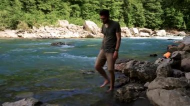 Sakallı ve rastalı yakışıklı beyaz adam soğuk dağ nehri kıyısında yalın ayak yürüyor ve hayatın tadını çıkarıyor. İnsan şortlu ve tişörtlü nehirde altını ıslatıyor. 4k görüntü.