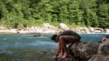 Sakallı ve rasta saçlı yakışıklı beyaz adam büyük bir taşın üzerinde oturuyor ve ayaklarını serin, fırtınalı bir dağ nehri mavisi renginde yıkıyor. Erkek turist saha koşullarında hijyen yapıyor..