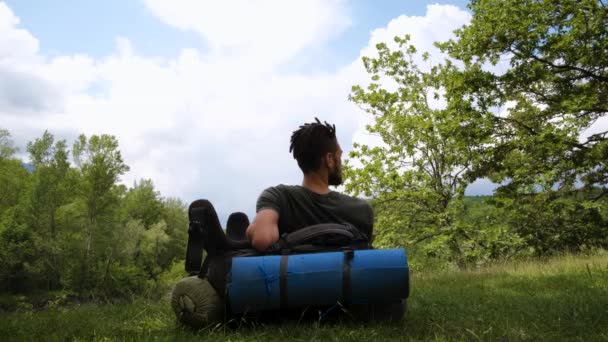 世界を探索し 人生を楽しむ ドレッドロックを持つ若いハンサムな白人男性は 大規模な観光バックパックにもたれ 彼の腕を伸ばして自然の中に座っている 一人旅 短い4K映像 — ストック動画