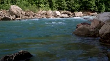 Yatay yakınlaştırma 4K yavaş çekim videosu. Mavimsi renkte güzel, fırtınalı bir dağ nehri büyük taşların arasında. Arka planda yoğun yeşil yaprak döken orman.