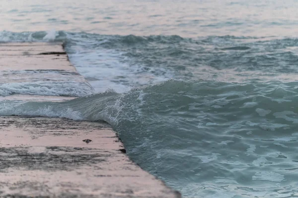 Blue sea wave rolls water on concrete pier. Pink reflection and glitter on pier from gentle dawn sky. Sea morning landscape clear transparent wave close up.