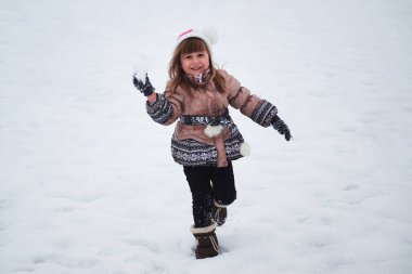 Küçük şirin beyaz kız kış parkında yürüyor ve kartopu atıyor. Çocuk örgü şapka takıyor ve kalın ceket giyiyor. Eğleniyor ve kış tatillerini doğada geçiriyor..