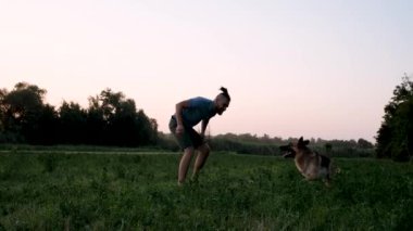 Saçları rasta desenli beyaz bir adam yazın günbatımında tarlada koşuyor ve Alman çoban köpeğiyle oynarken eğleniyor. Köpekle aktif ve enerjik bir yürüyüş. Yavaş çekim 4k video.
