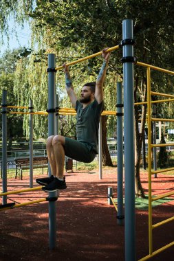 Genç yakışıklı beyaz adam sıcak bir yaz sabahı kendini sokak sporları sahasına atıyor. Kol kasları için dayanıklılık eğitimi. Aktif yaşam tarzı konsepti. Yatay çubuğa tutun ve bacaklarını kaldır.