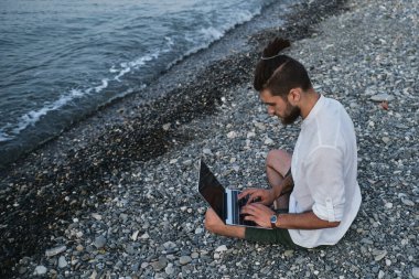 Hippi işadamı seyahat üzerine çalışır ve internette blog yazar. Başarılı beyaz erkek serbest yazar plajda oturuyor ve uzaktan bilgisayarla çevrimiçi çalışıyor. Dışarıdaki işe odaklan..