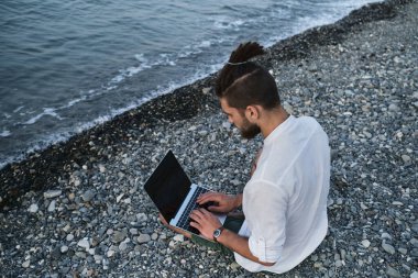 Hippi işadamı seyahat üzerine çalışır ve internette blog yazar. Başarılı beyaz erkek serbest yazar plajda oturuyor ve uzaktan bilgisayarla çevrimiçi çalışıyor. Dışarıdaki işe odaklan..