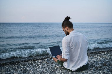 Hippi işadamı seyahat üzerine çalışır ve internette blog yazar. Başarılı beyaz erkek serbest yazar plajda oturuyor ve uzaktan bilgisayarla çevrimiçi çalışıyor. Dışarıdaki işe odaklan..