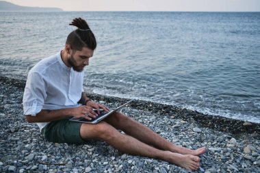 Hippi işadamı seyahat üzerine çalışır ve internette blog yazar. Başarılı beyaz erkek serbest yazar plajda oturuyor ve uzaktan bilgisayarla çevrimiçi çalışıyor. Dışarıdaki işe odaklan..