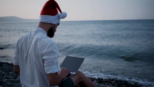 Giovane Freelance Rosso Cappello Babbo Natale Siede Sulla Spiaggia Ghiaia — Video Stock