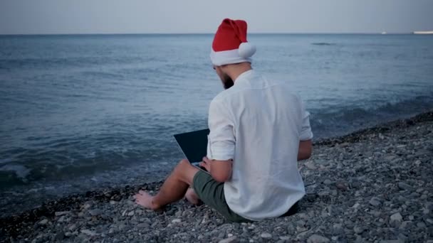 Homem Jovem Freelance Chapéu Vermelho Papai Noel Senta Praia Seixos — Vídeo de Stock