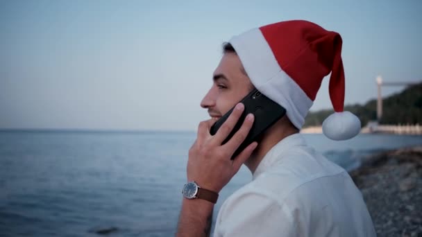 お正月とクリスマスをお祝いしましょう 白いシャツを着た若いフリーランスの従業員と 海のそばに座って電話で話している赤いサンタの帽子 遠隔地での旅行やオンライン作業を組み合わせる — ストック動画