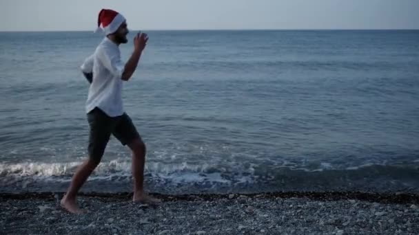 4Kスローモーションビデオ 白いシャツを着た若い白人男性と赤いサンタの帽子は 青い海の背景を背景に小石のビーチに沿って単に走ります 新年とクリスマスを創造的に祝う — ストック動画