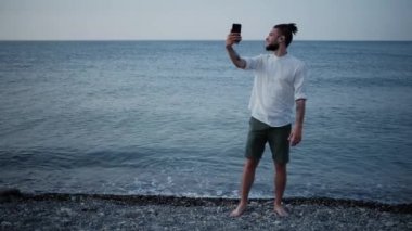 Sakallı, beyaz tişörtlü, kulaklıklı, plajda kulaklık takan ve telefonda sohbet eden beyaz bir genç. Uzaktan çalışma ve tatilde iletişim. Başarılı iş adamı.