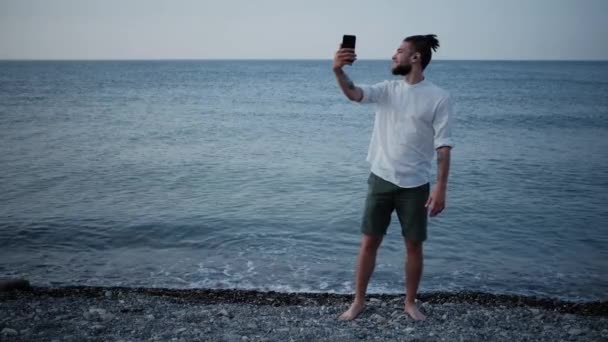 수염을 기르고 셔츠를 남성은 해변에서 이어폰을 통화를 실시간으로 채팅을 휴가중인 — 비디오