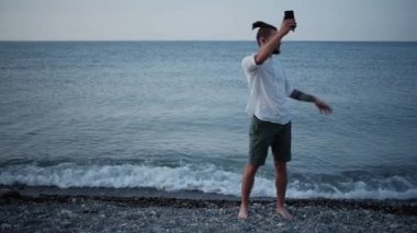 Sakallı, beyaz tişörtlü, kulaklıklı, plajda kulaklık takan ve telefonda sohbet eden beyaz bir genç. Uzaktan çalışma ve tatilde iletişim. Başarılı iş adamı.