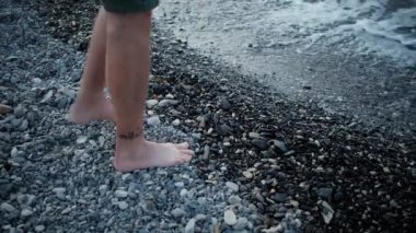 Kayalık kıyı boyunca yürür ve ayak bileklerine kadar denize açılır. Temiz deniz suyunda ıslak ayaklar. 4K yavaş çekim videosu suyun içindeki ayakları yakından gösteriyor..